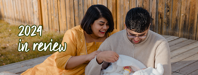 text: 2024 in review. a picture of woman (Aurooba) in a yellow dress leaning over a smiling man's (Aurooba's spouse) shoulder to peer at a child wrapped in a white fuzzy blanket in his arms, with a smile. The family is sitting on a wooden deck and you can see a yellow fence and some grass in the background.